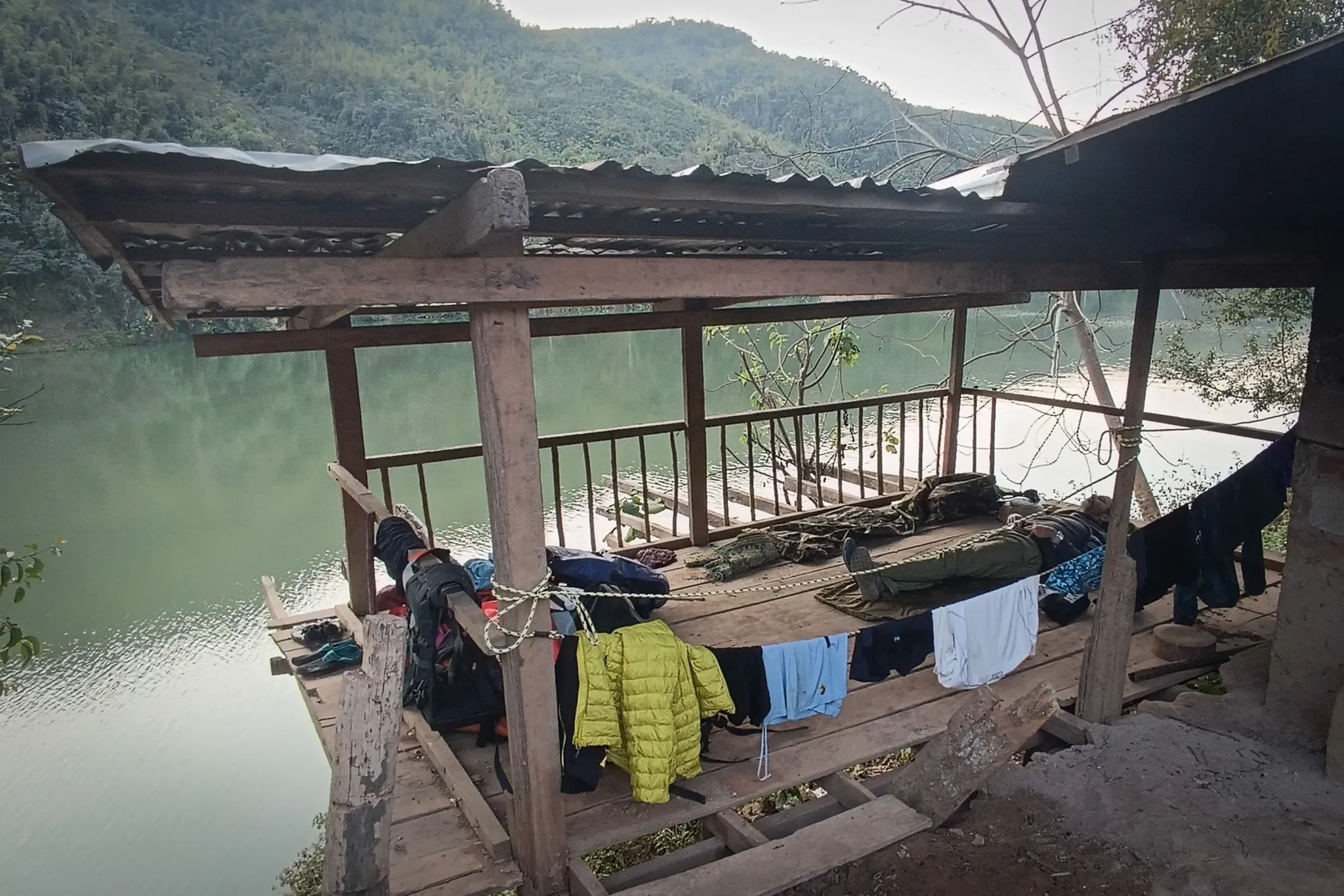 Exterior view of the raised open shelter used as a camp with clothes drying and the calm Nam Ou river and forested hills behind