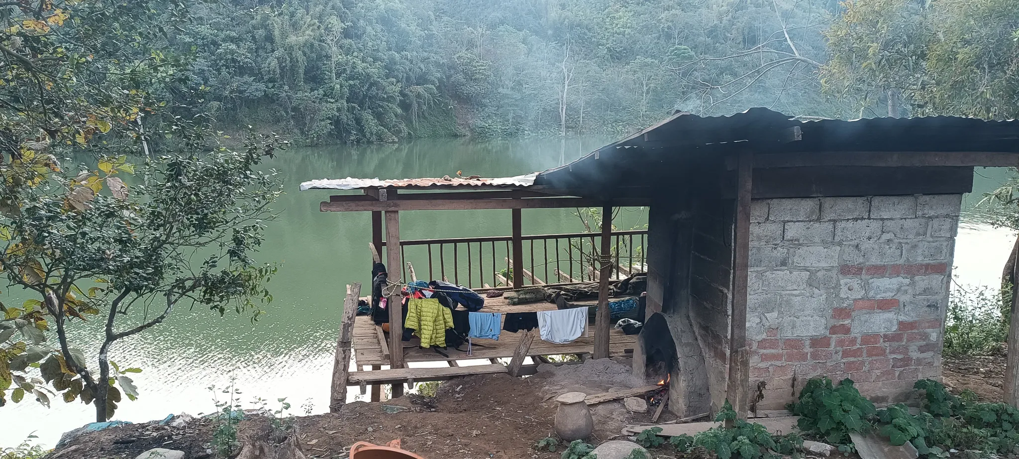 Open riverside shelter with clothes drying on a line and a brick oven with smoke rising, overlooking the misty Nam Ou river