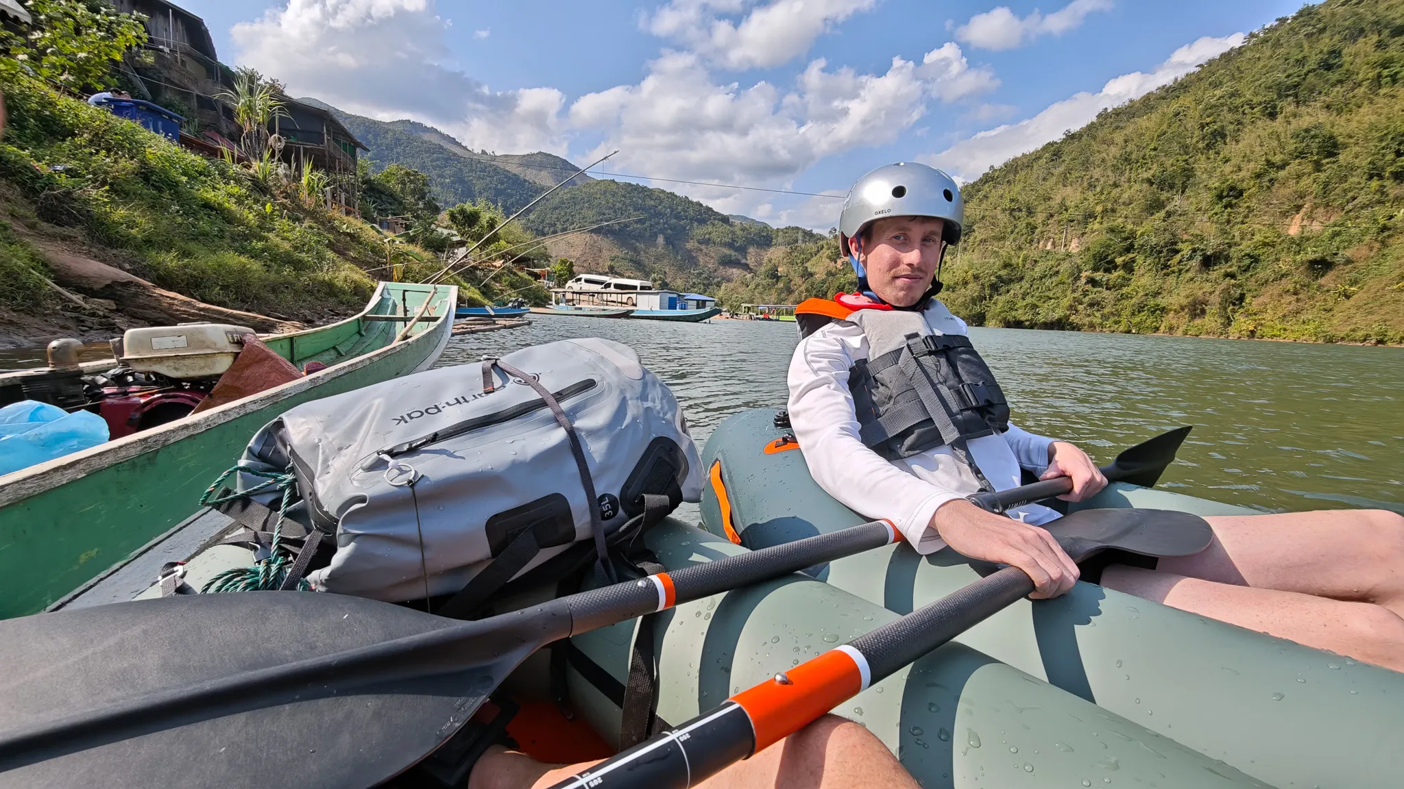 Packrafter paddling towards the suspension bridge on the Nam Ou river with Muang Khua town and palm trees on the hillside