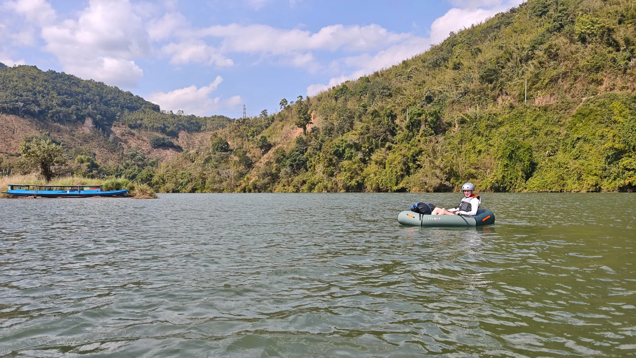 Distant view of a packrafter on the expansive Nam Ou river near Muang Khua, surrounded by green mountain hillsides