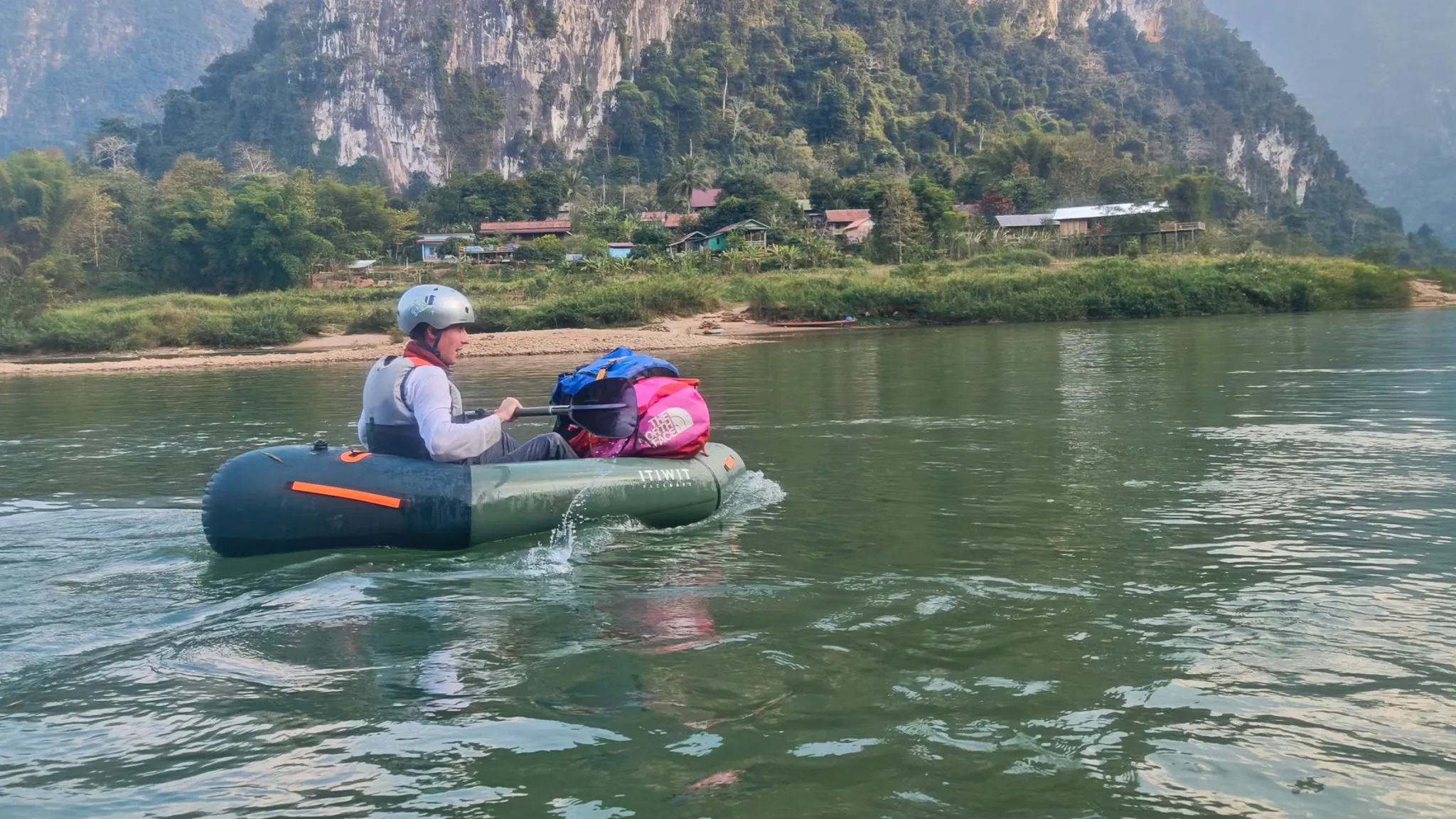 Packrafter in helmet and buoyancy aid paddling a loaded Itiwit kayak towards Muang Ngoi with towering karst mountains behind