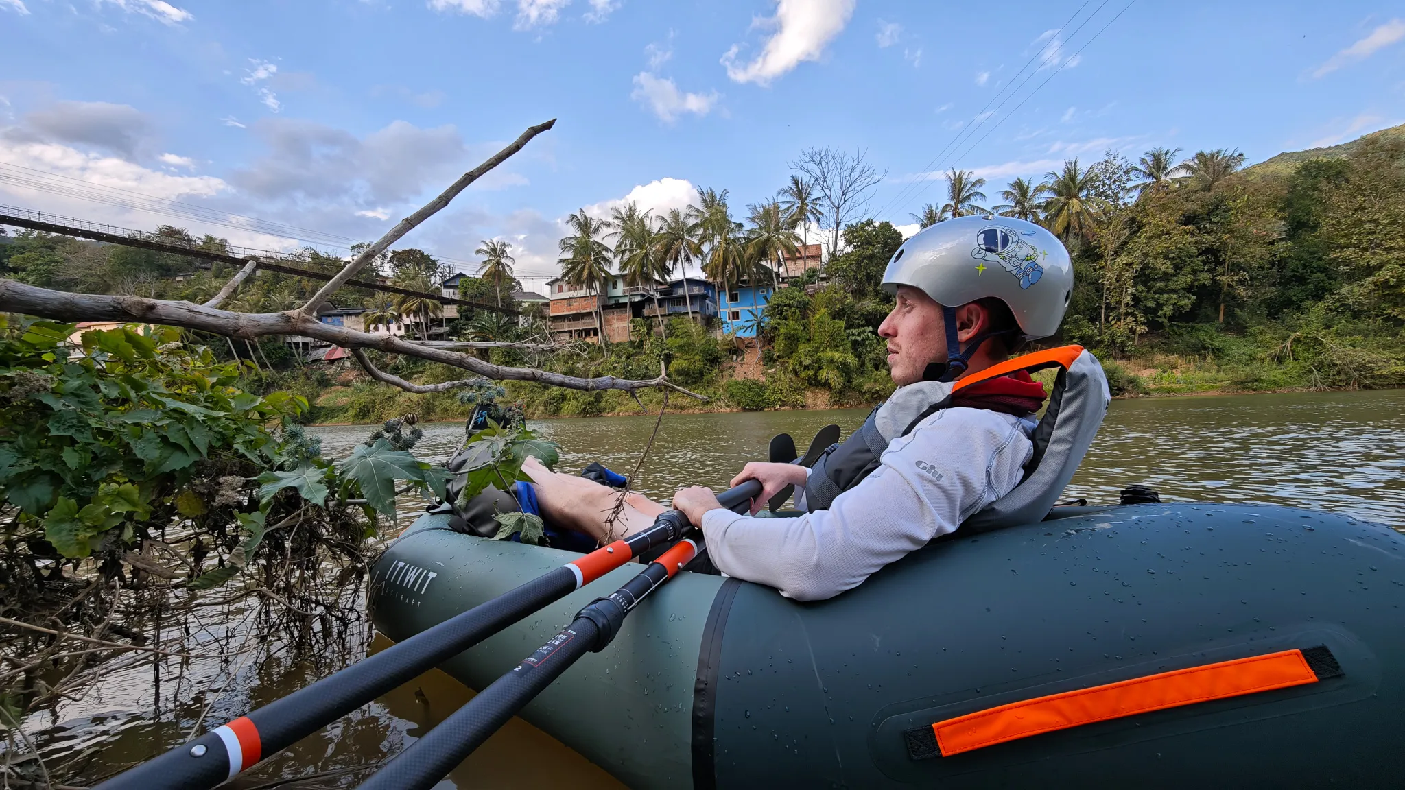 Packrafter lying back in an Itiwit raft drifting near the riverbank on the Nam Ou at Muang Khua with the suspension bridge behind