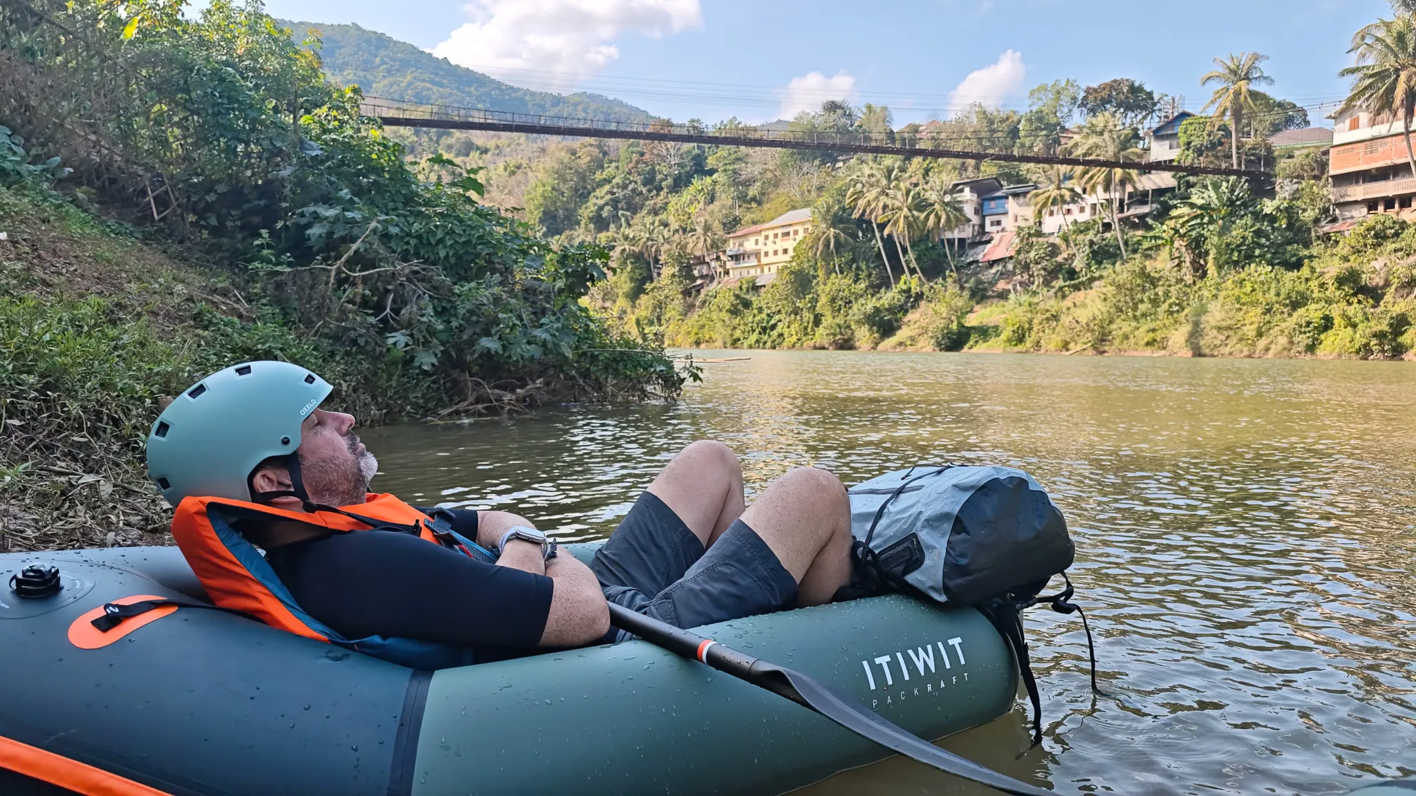Packrafter leaning back looking at the sky in an Itiwit raft beneath the Muang Khua suspension bridge on the Nam Ou river