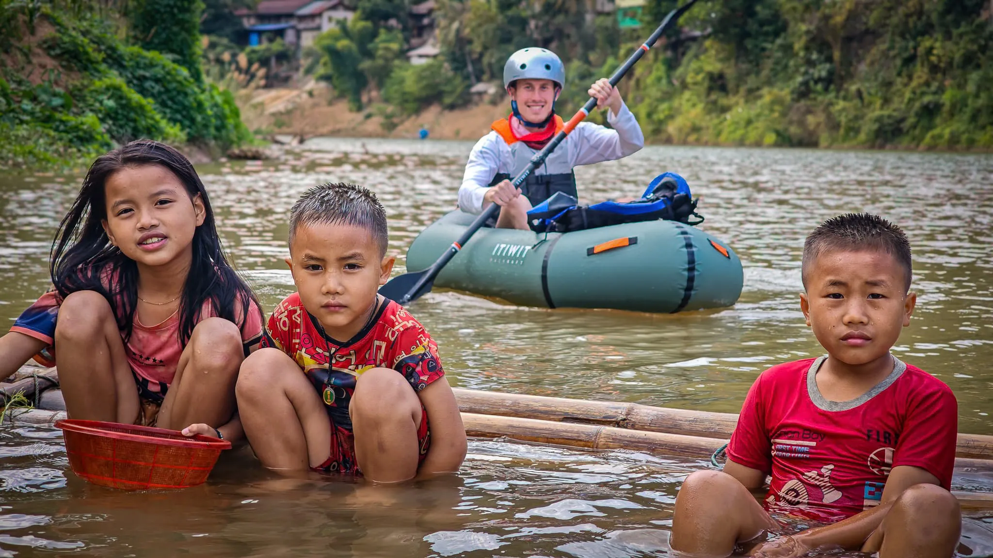 Packrafter in Itiwit raft paddling near the riverbank with Muang Khua town and suspension bridge visible in the background