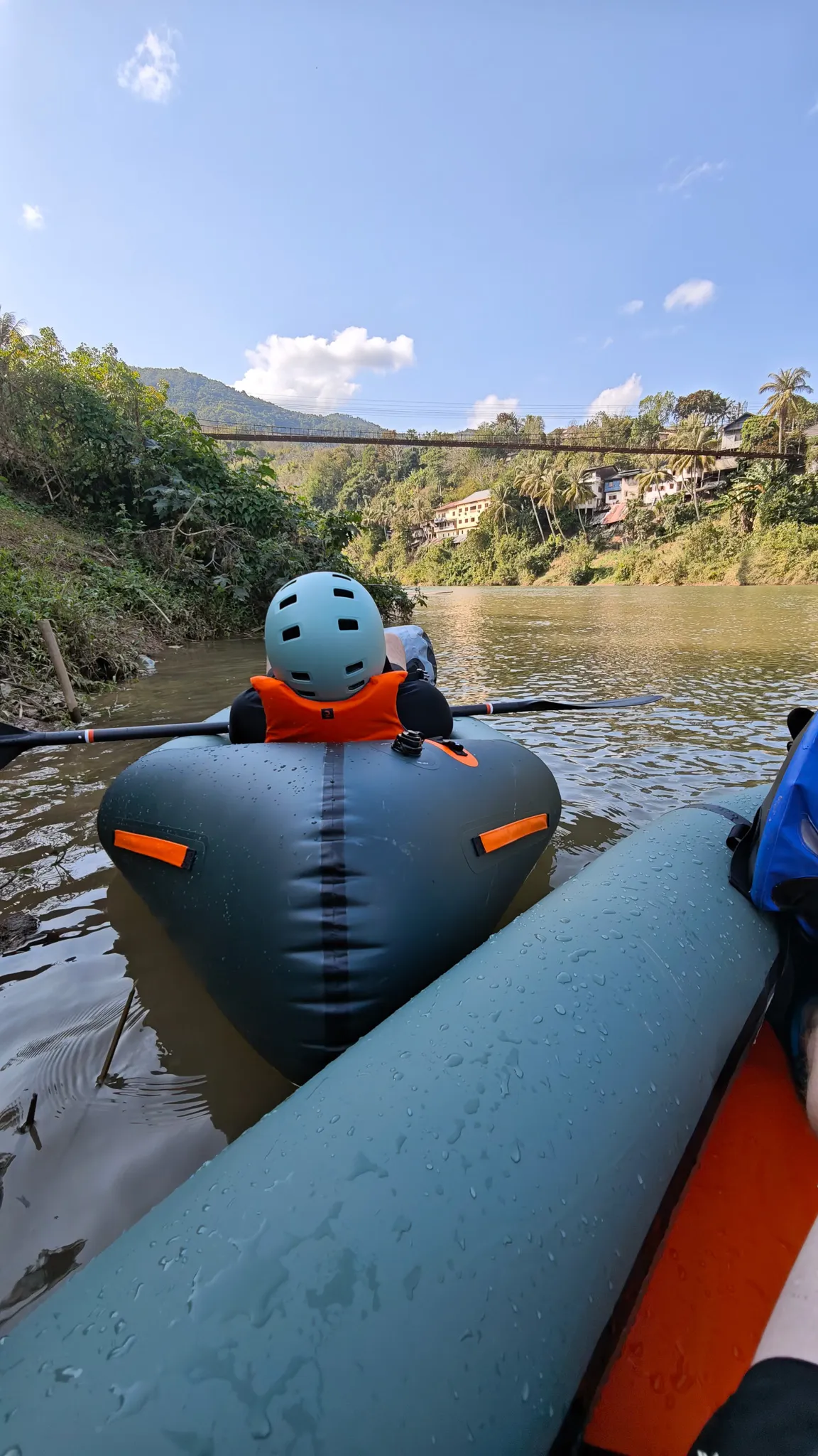 Packrafter sitting in an Itiwit raft near fallen tree branches on the Nam Ou river with the suspension bridge and Muang Khua behind