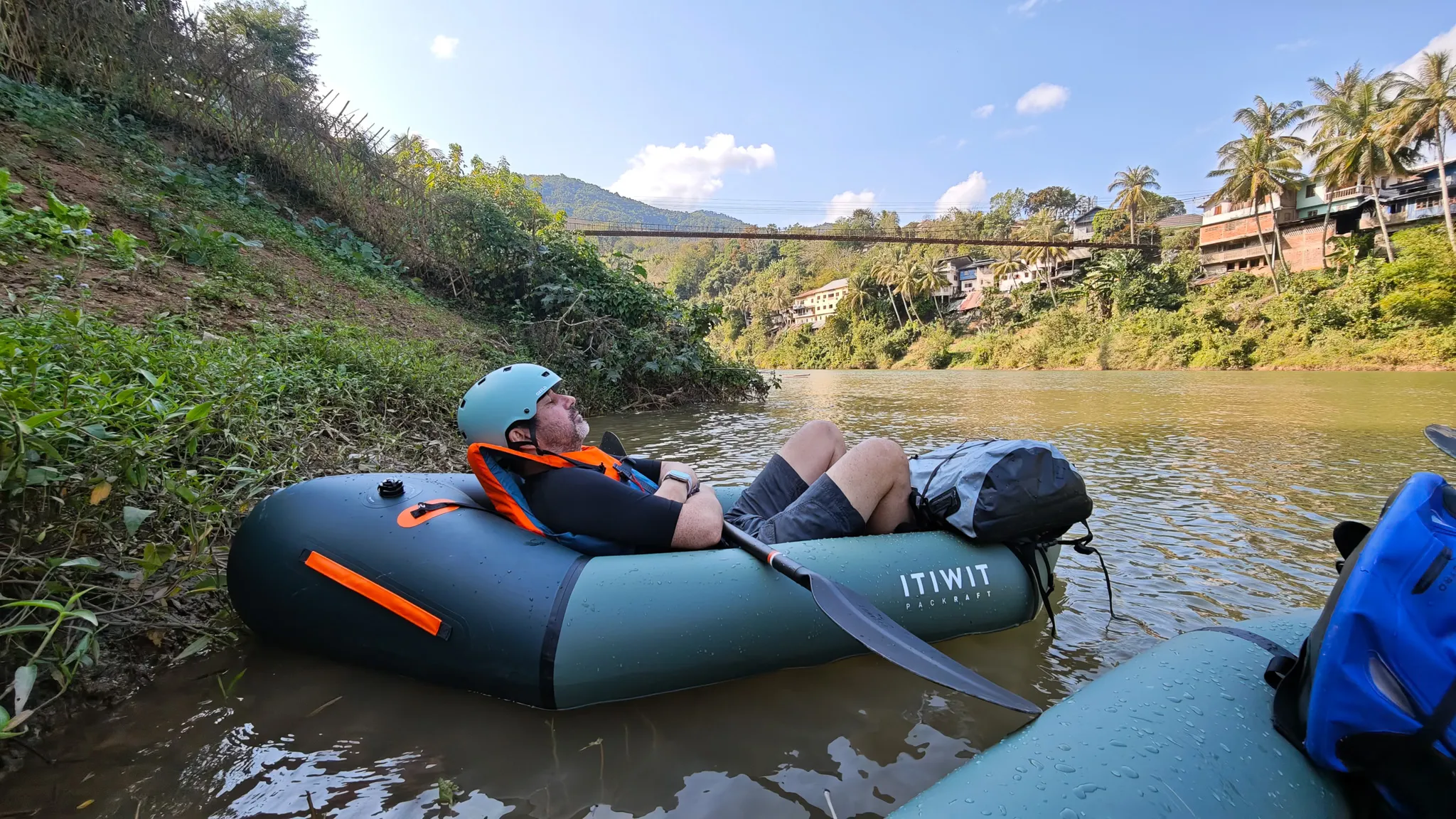 Packrafter reclining in an Itiwit raft on the Nam Ou river with the suspension bridge and Muang Khua hillside town visible