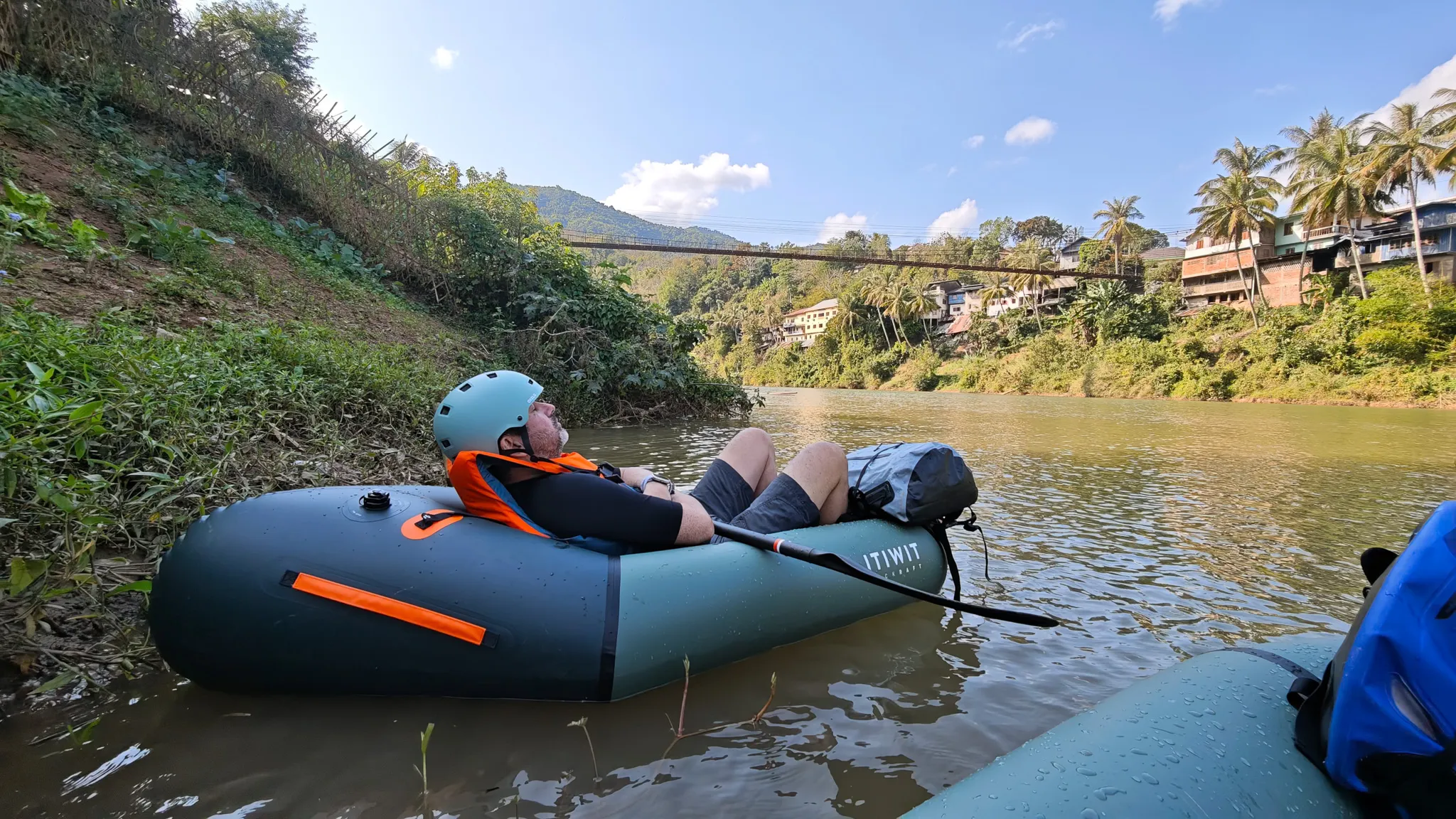 Relaxed packrafter resting in an Itiwit raft on the Nam Ou with the suspension bridge and palm trees framing the scene at Muang Khua