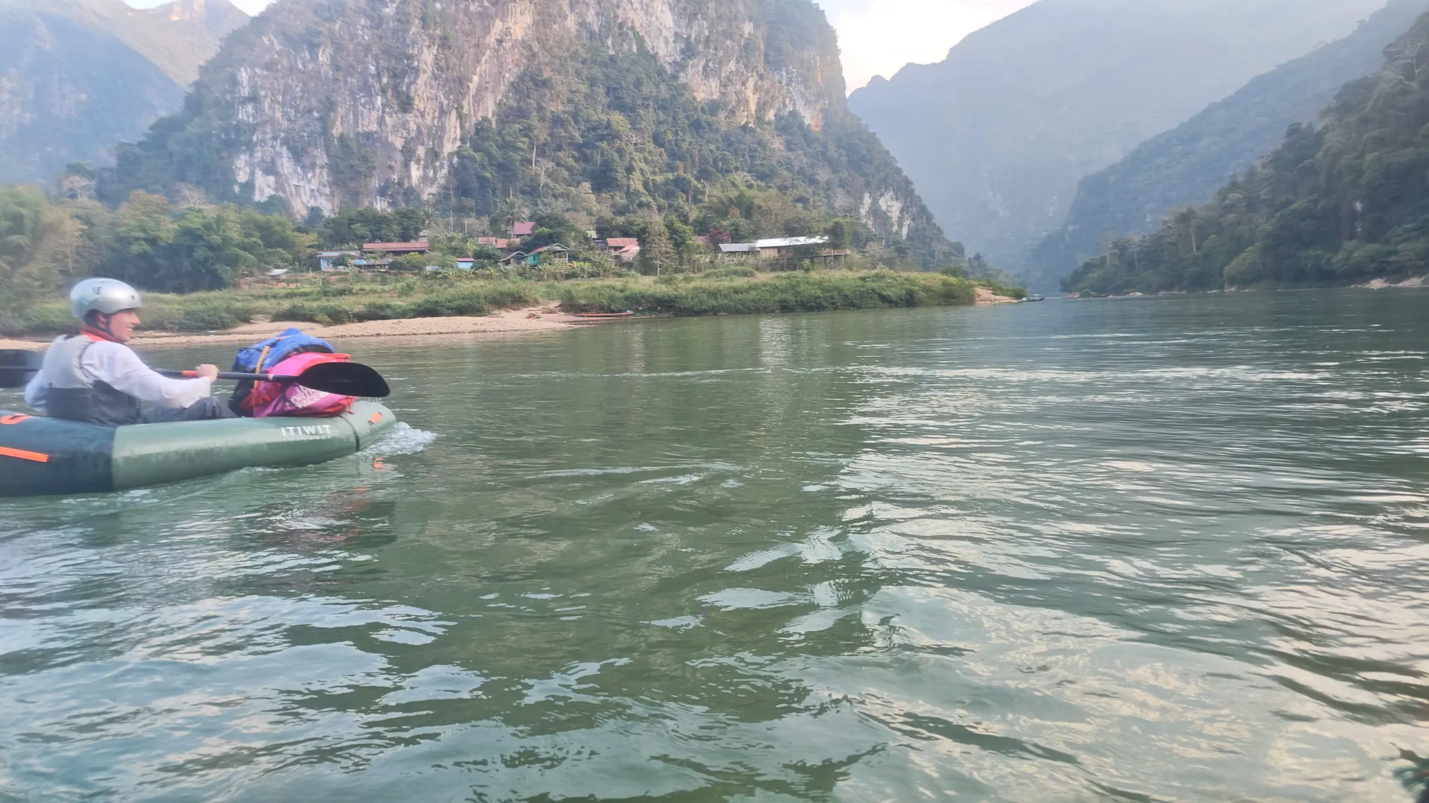 Packrafter paddling an Itiwit kayak on the Nam Ou river with dramatic karst limestone cliffs and Muang Ngoi village ahead