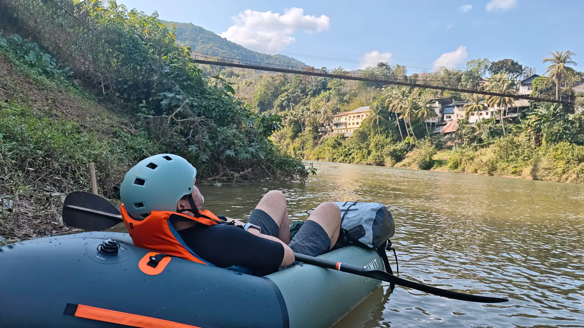 Two packrafts drifting under the suspension bridge on the Nam Ou river with Muang Khua town and blue sky overhead