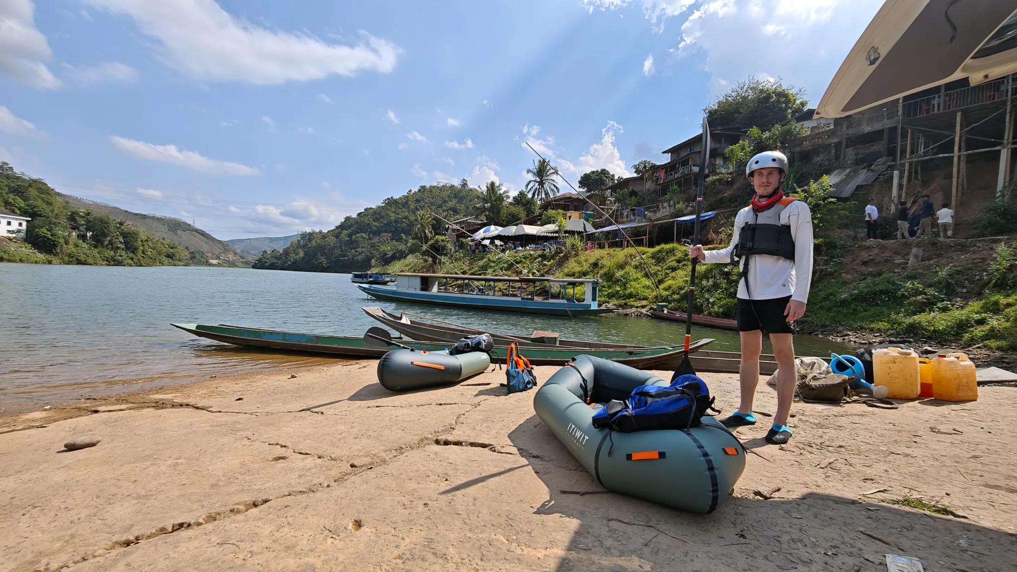 Traveller in helmet and life jacket standing beside inflated Itiwit packrafts on the sandy bank of the Nam Ou river, ready to launch at Muang Khua