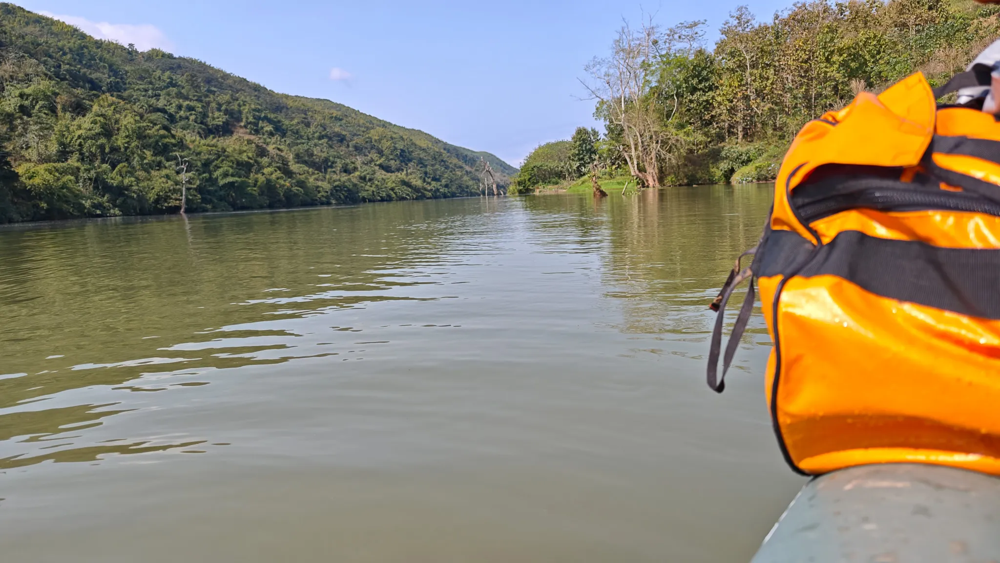 Paddling downstream on the Nam Ou river through a forested valley with an orange dry bag visible on the packraft bow