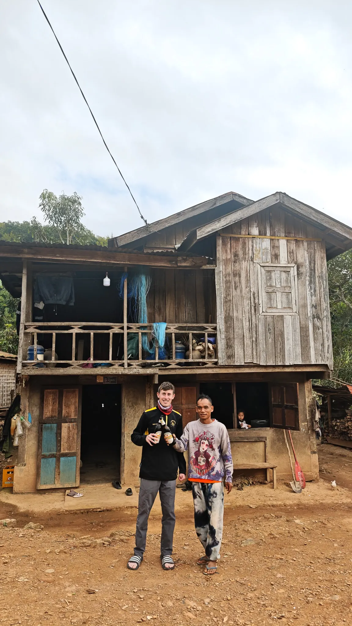 Portrait of a traveller and his Lao host standing together with Beer Lao in front of a traditional wooden stilt house in Ban Haddean