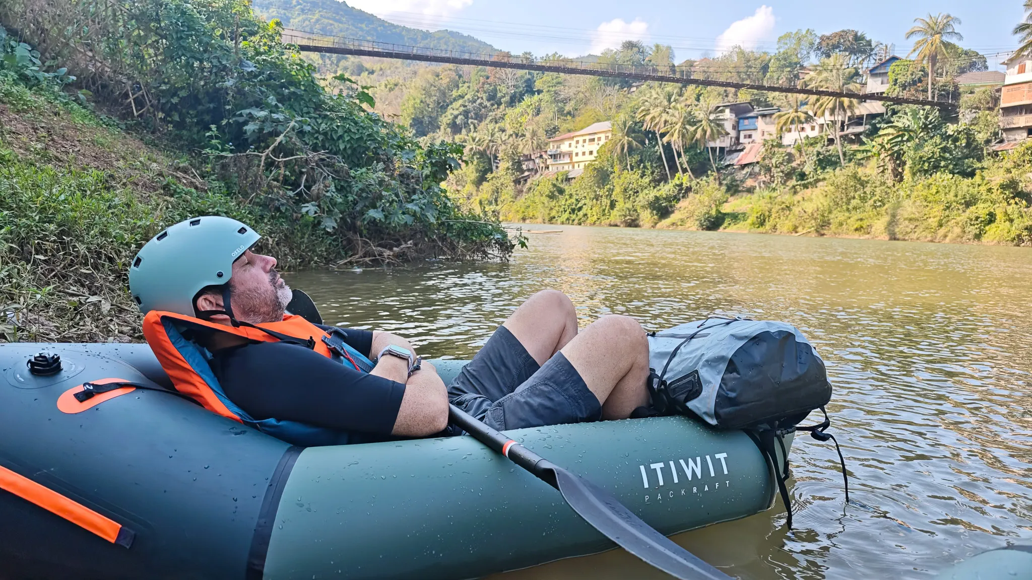 Paddler lying back in a packraft on the calm Nam Ou river with the Muang Khua suspension bridge and tropical vegetation behind