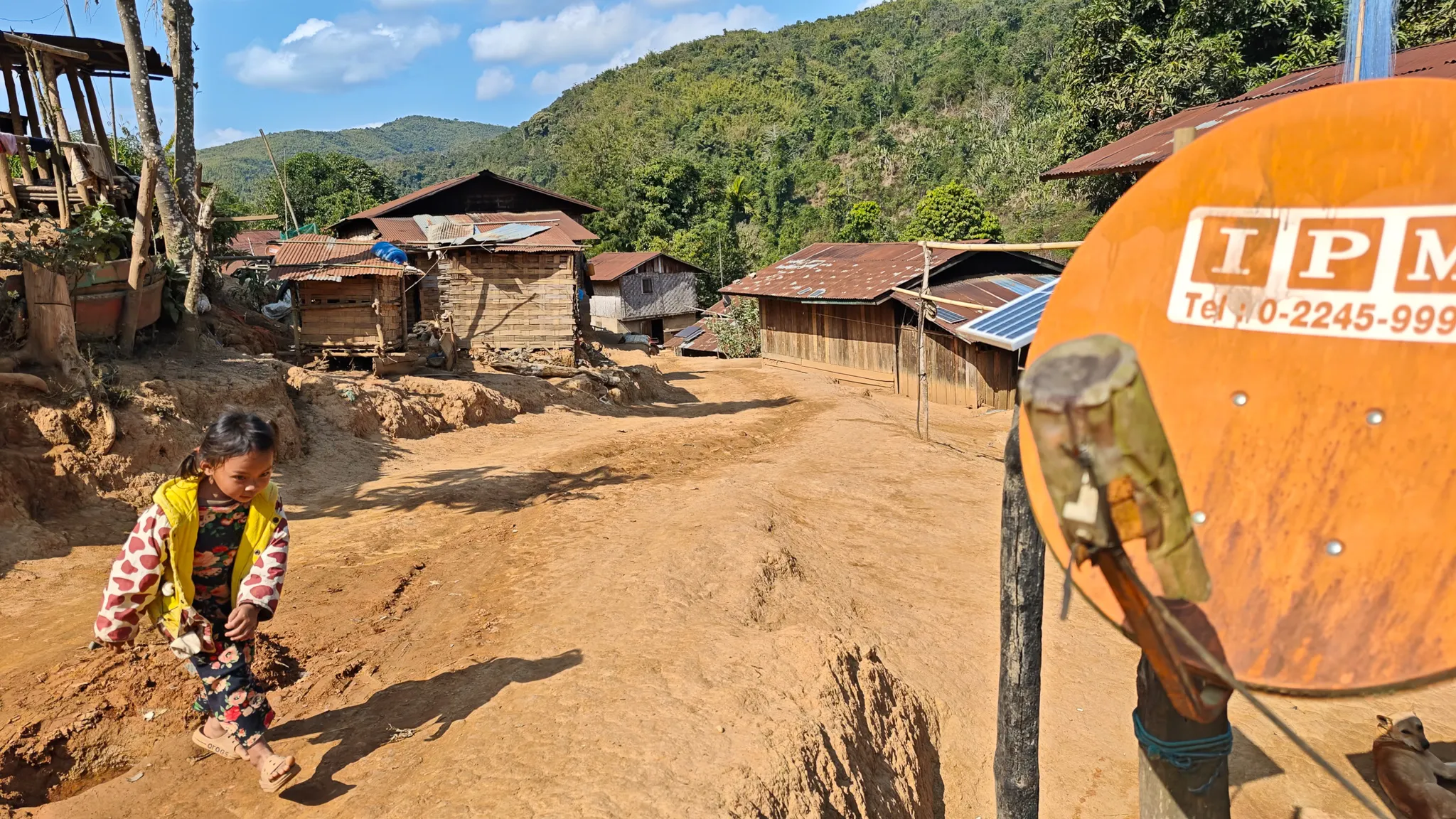 A remote Lao village with wooden houses, a satellite dish, and a child on a dusty road near the Nam Ou river
