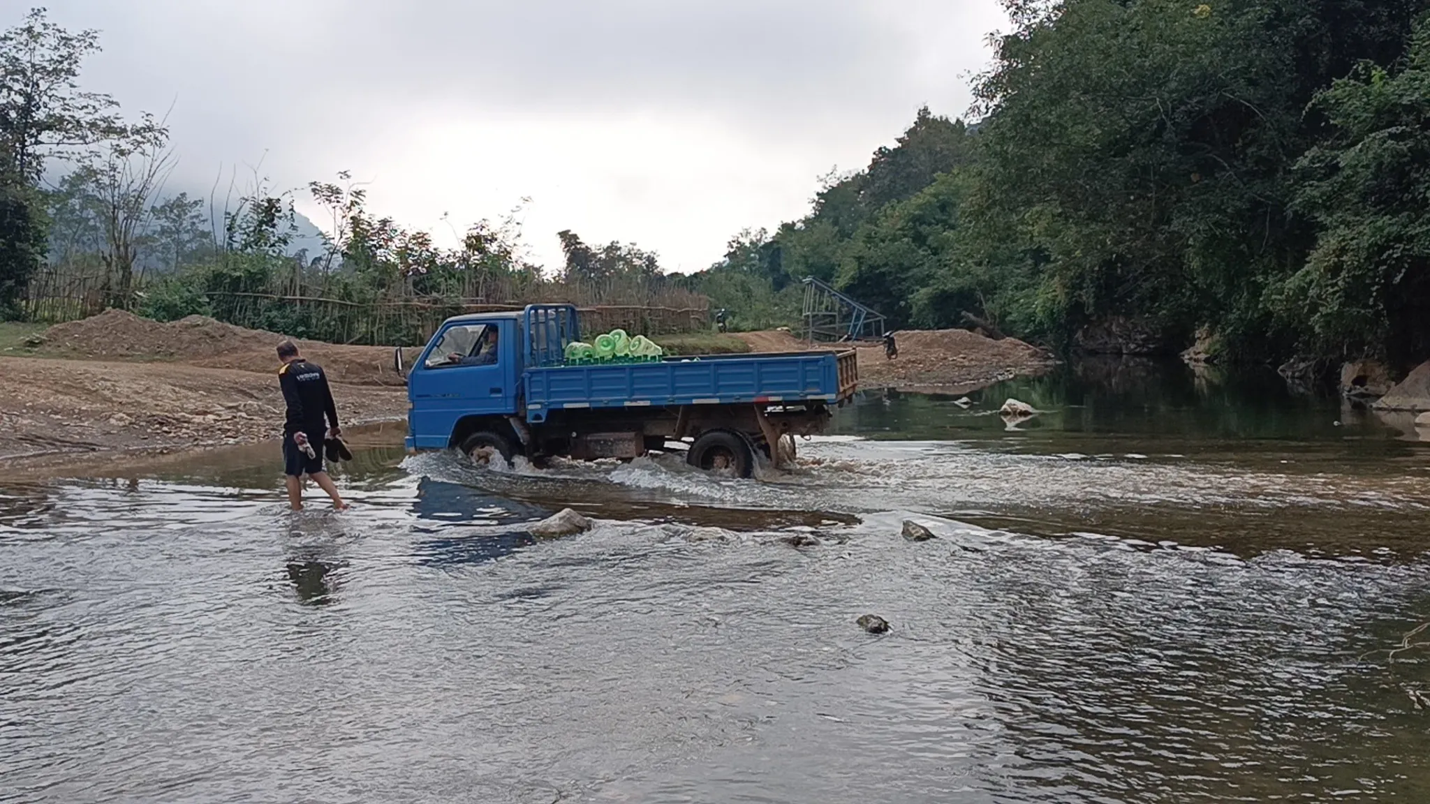Man wading across a river ford alongside a blue truck near Muang Ngoi during the hike back from the cave