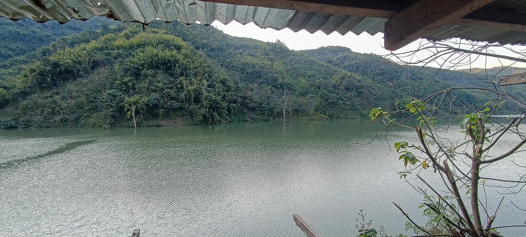The Nam Ou river and forested hills framed by the tin roof of an abandoned shelter used as a camp on the riverbank