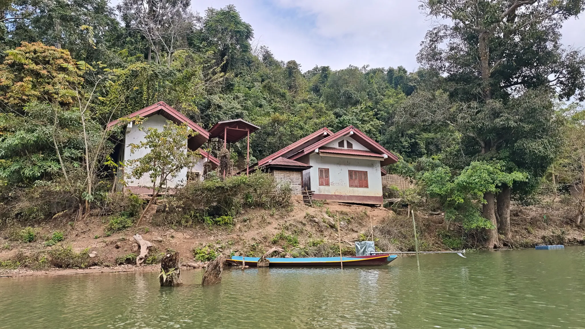 A riverside village seen from the water with stilted houses on a steep bank and a blue longboat moored below on the Nam Ou river