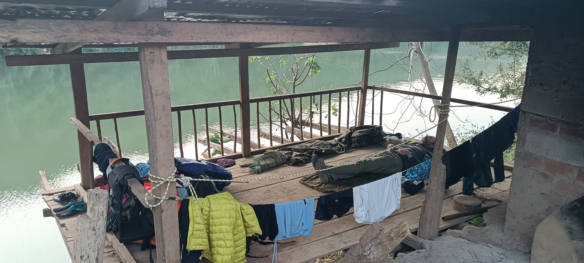 Sleeping setup on the wooden floor of an open shelter with gear and clothes drying, overlooking the green Nam Ou river