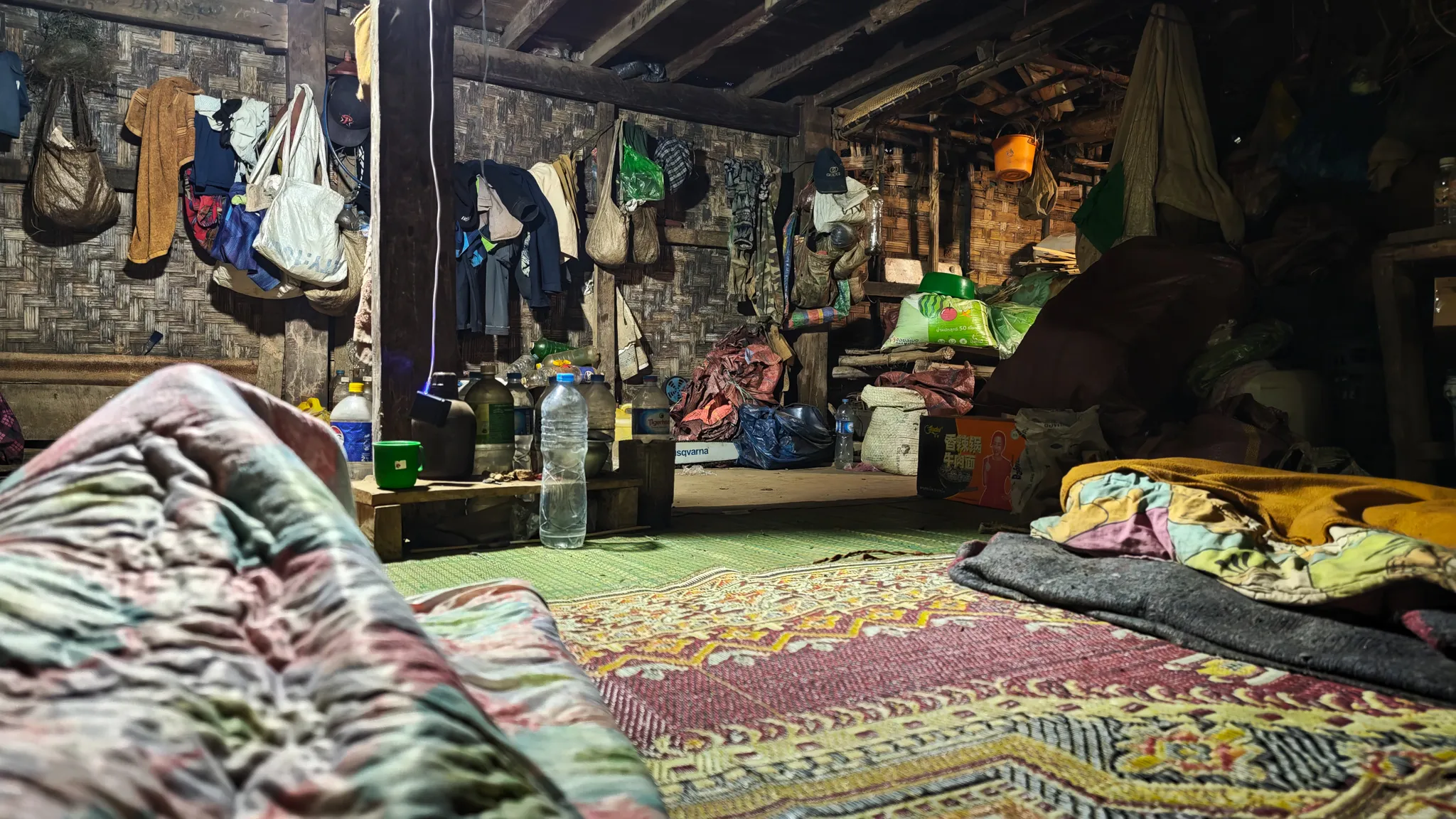 Interior of a traditional village hut with woven bamboo walls, sleeping mats, and bags hanging from the rafters along the Nam Ou river in Laos