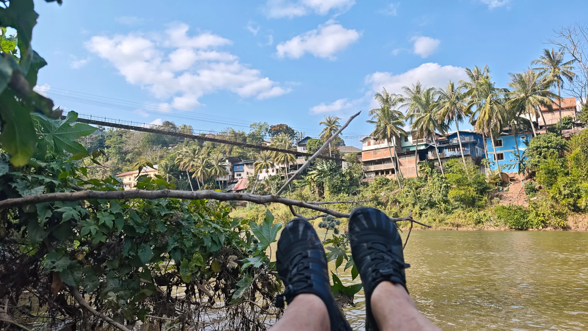 Harry paddling an Itiwit packraft on the Nam Ou river near Muang Khua with the town and suspension bridge visible beyond palm trees