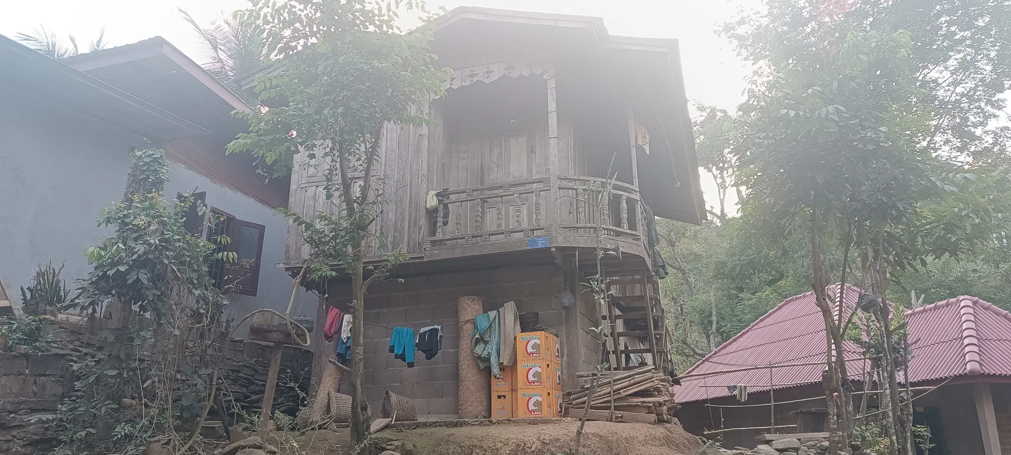Two-storey traditional Lao stilt house with ornate wooden balcony and laundry hanging below in Ban Haddean village