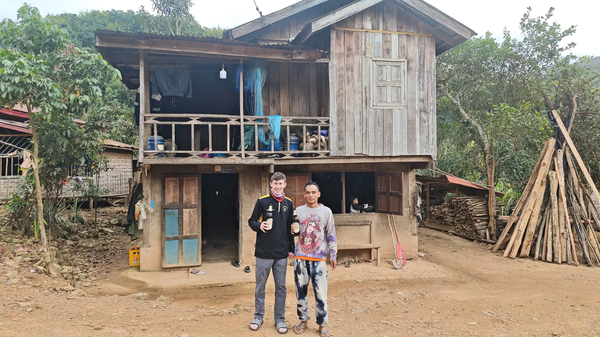 Traveller and local host holding Beer Lao bottles pose in front of a two-storey wooden stilt house in Ban Haddean village