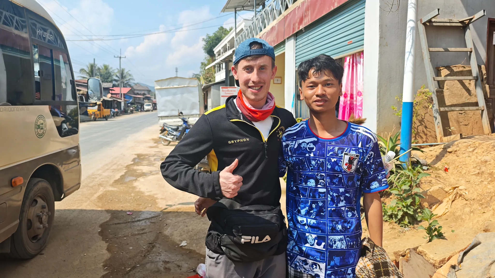 Traveller posing with thumbs up alongside a friendly local in a Japanese football shirt at a bus stop en route to Muang Khua