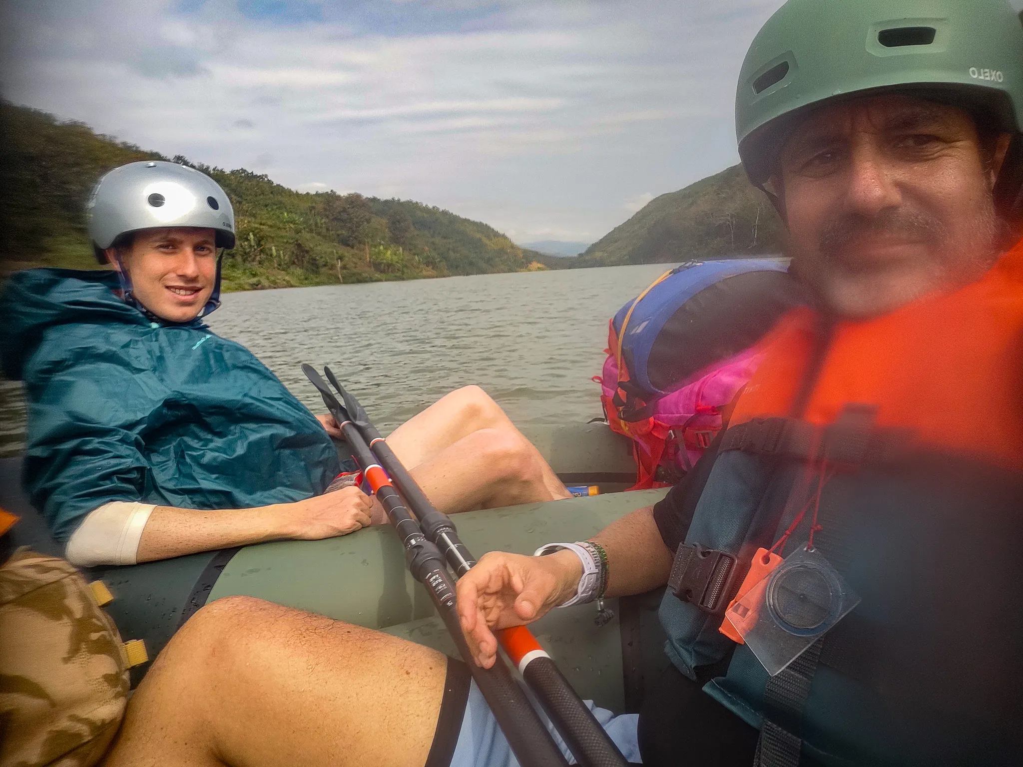 Two packrafters in helmets and life jackets taking a selfie in their inflatable kayak on the calm Nam Ou river surrounded by green hills