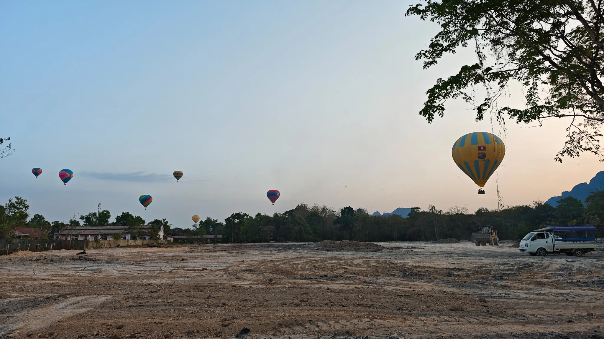 Multiple hot air balloons preparing to land in a dusty field at sunset with karst mountains in Vang Vieng