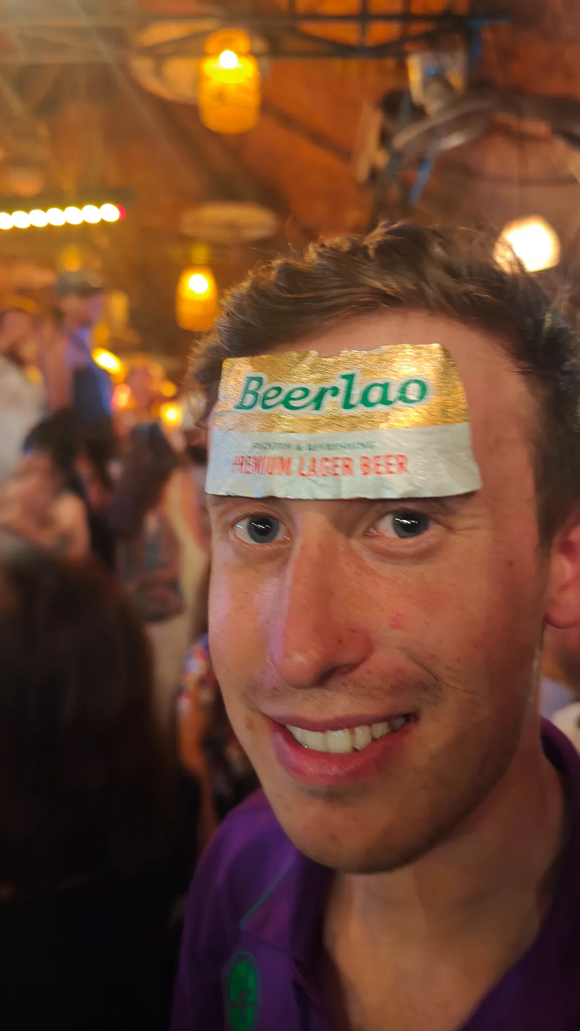 Close-up selfie with a Beer Lao label on forehead at a lively bar during the Vang Vieng night out