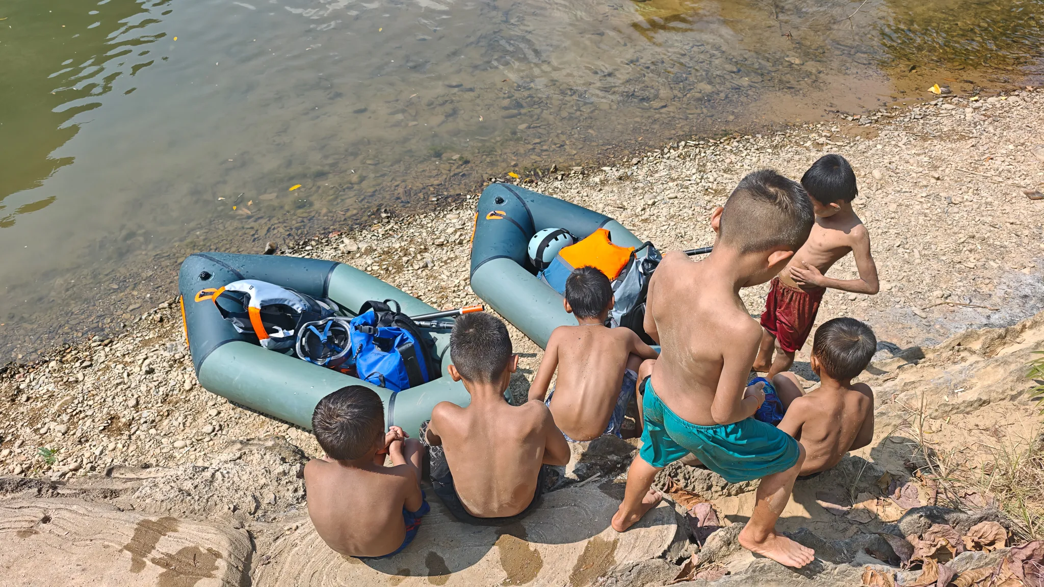 Lao children sitting on rocks examining an Itiwit packraft beached on the shore of the Nam Song River