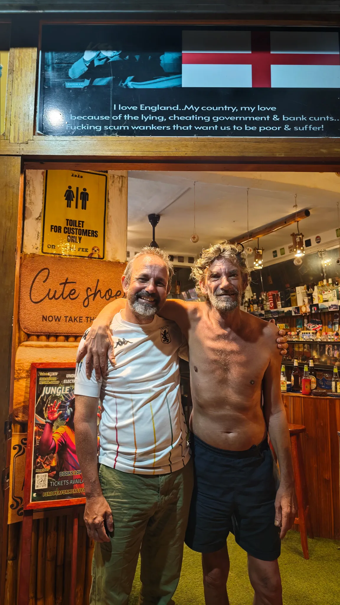 Two friends in the doorway of a Vang Vieng dive bar with an England flag and cute shoes doormat sign