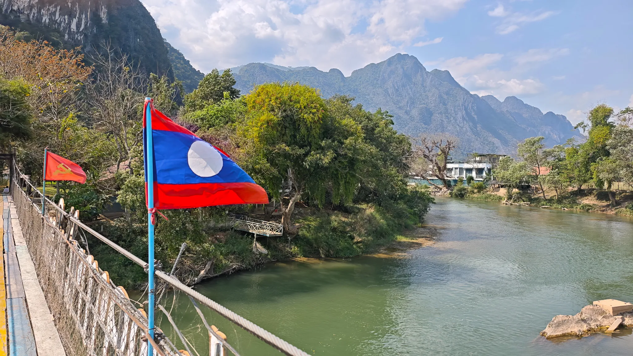 Laos flag flying from a suspension bridge with karst mountains and the Nam Song River in Vang Vieng