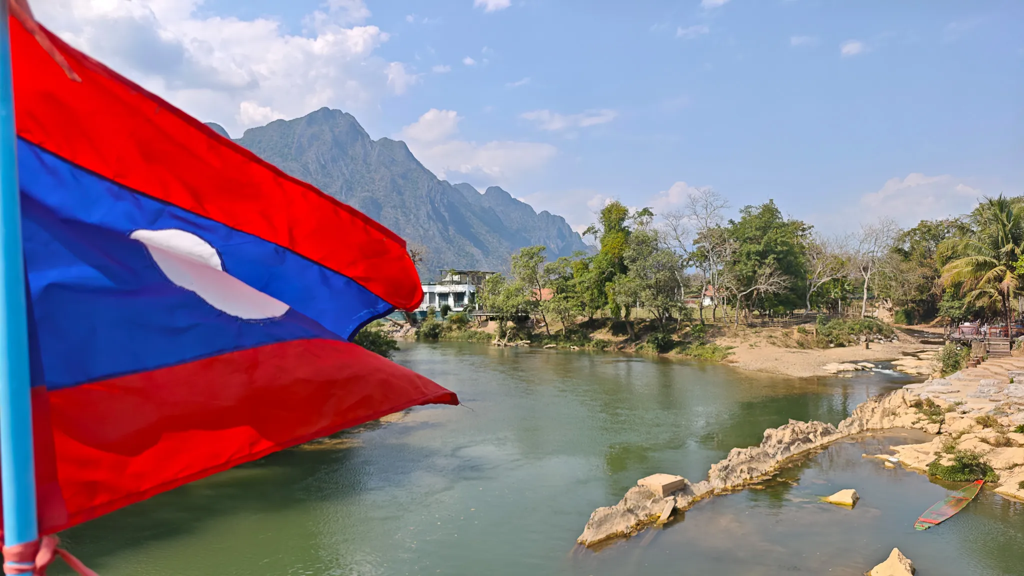 Laos flag flying over the Nam Song River with dramatic karst mountains and rocky shoreline in Vang Vieng