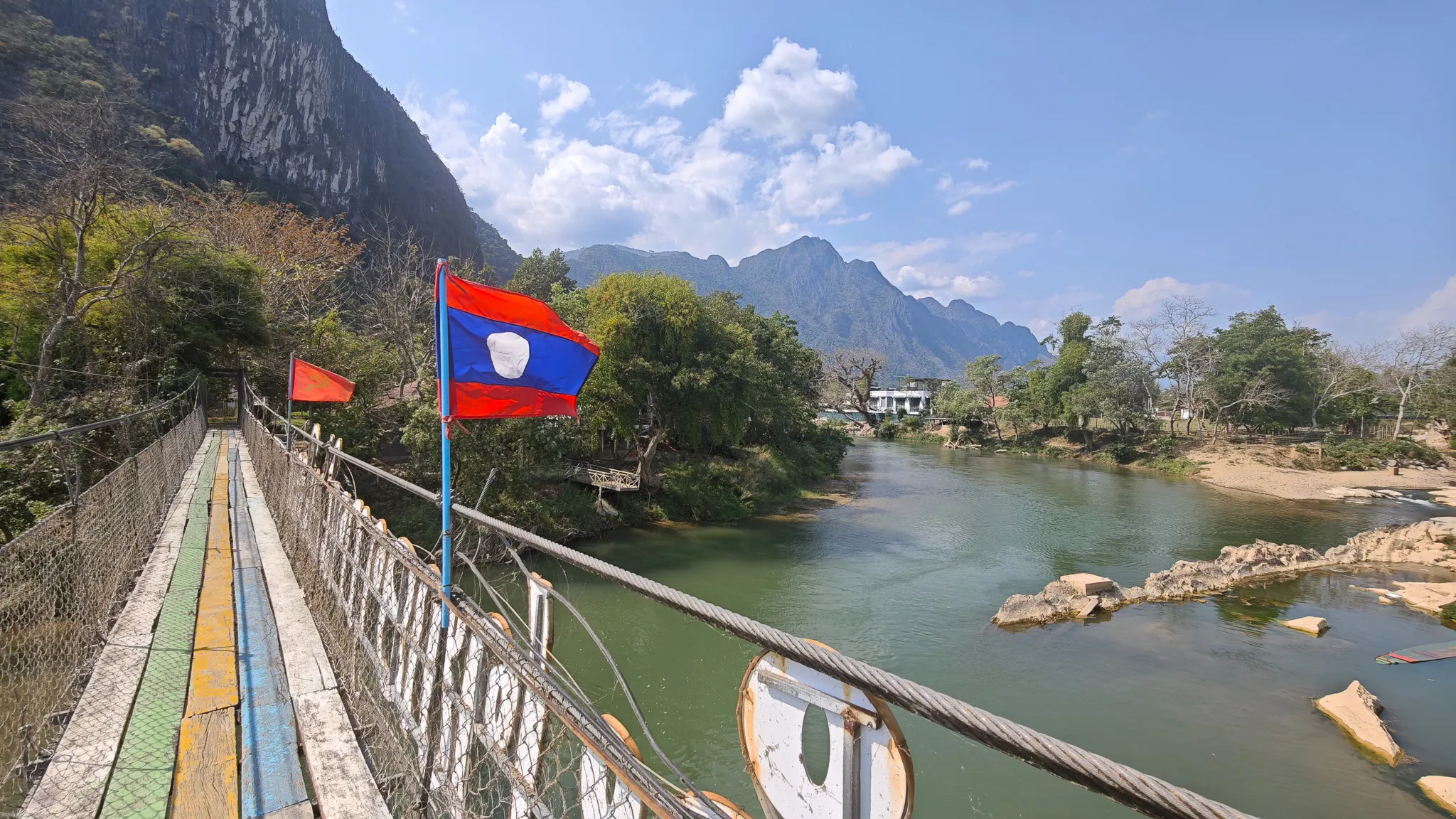 Laos and communist flags flying on a suspension bridge with panoramic views of karst mountains along the Nam Song River