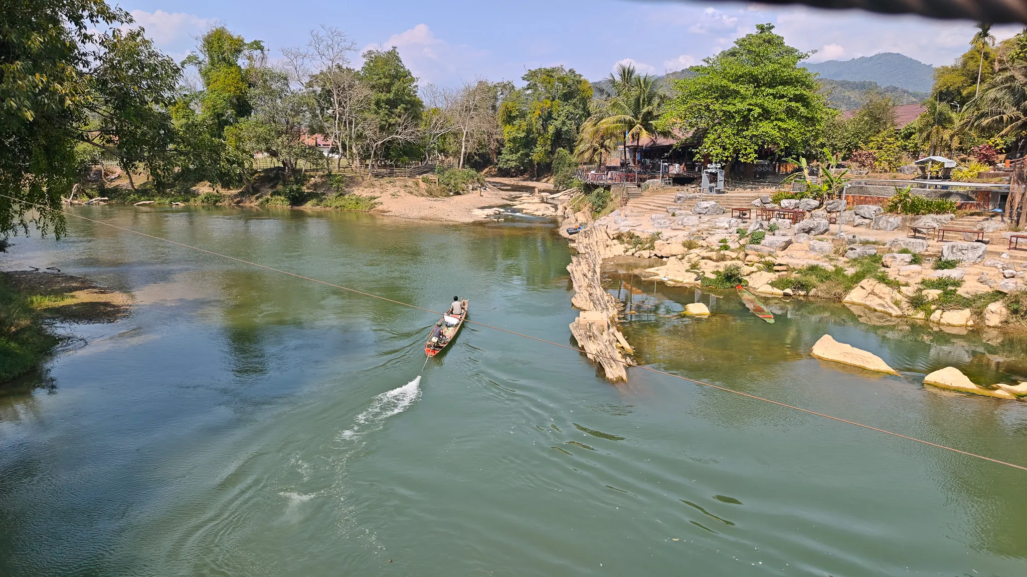 Traditional Lao longboat motoring through the Nam Song River viewed from a suspension bridge in Vang Vieng