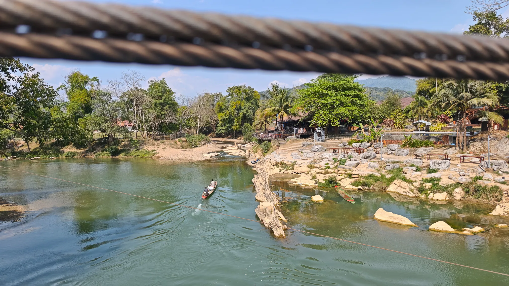 Longboat on the Nam Song River seen through steel bridge cables with rocky riverside bars in Vang Vieng