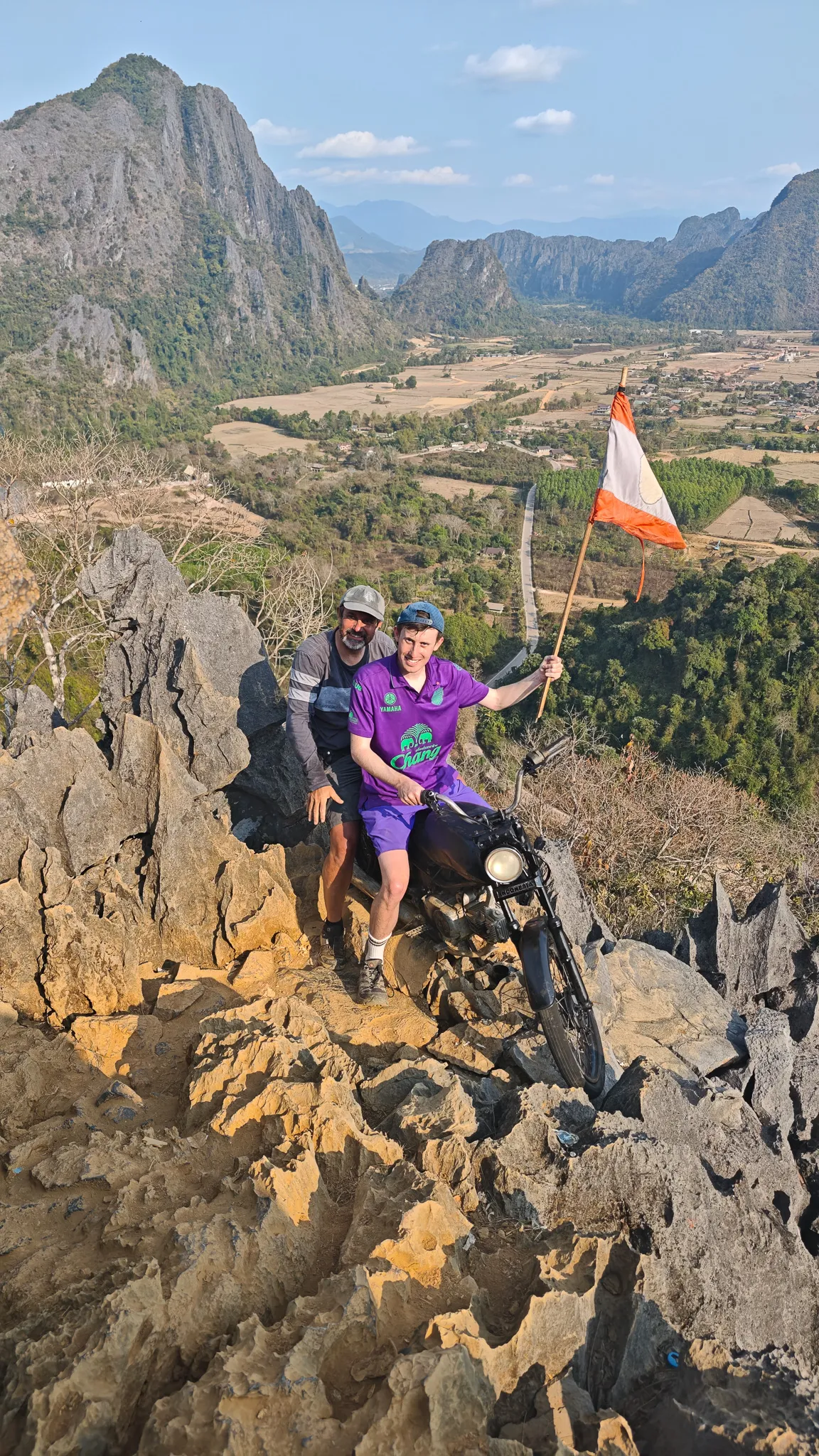 Sitting on a motorcycle with Laos flag at the Nam Xay viewpoint summit overlooking rice paddies and karst mountains