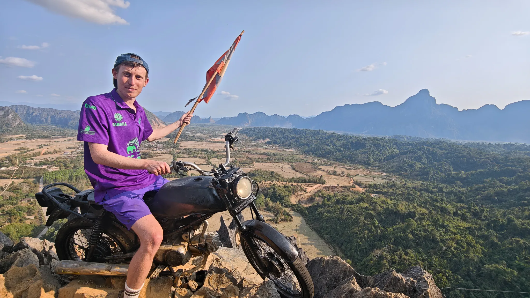 Wide panoramic shot of a person on a motorcycle at the Nam Xay summit with Laos flag and karst valley views