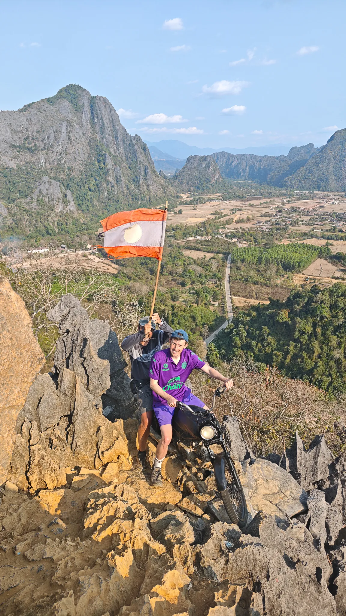 Holding the Laos flag at the Nam Xay viewpoint summit with dramatic karst mountain panorama