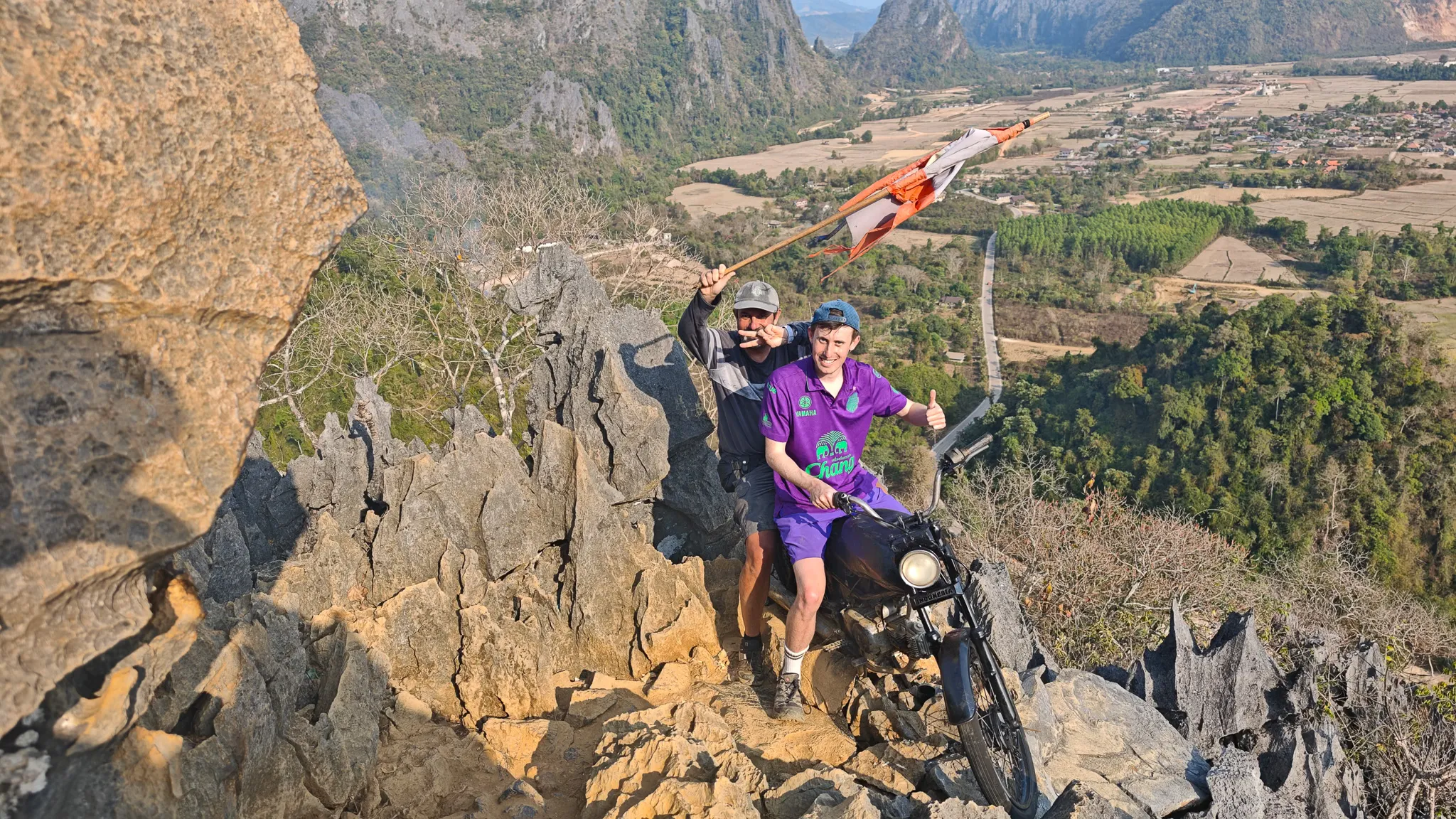 Pair posing with the Laos flag at Nam Xay viewpoint summit with dramatic karst backdrop