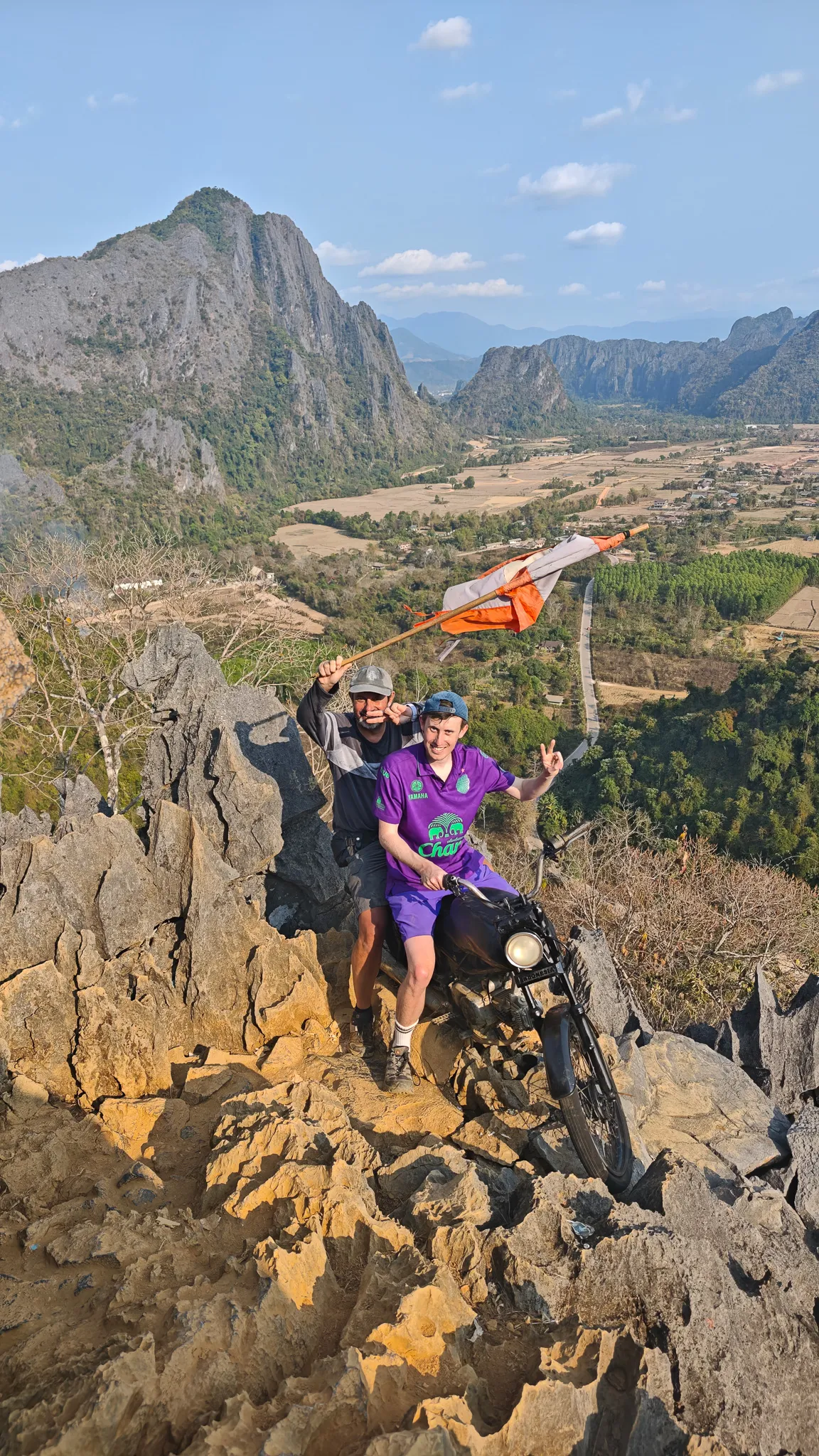 Wide shot of two friends with the Laos flag at the rocky peak of Nam Xay viewpoint in Vang Vieng