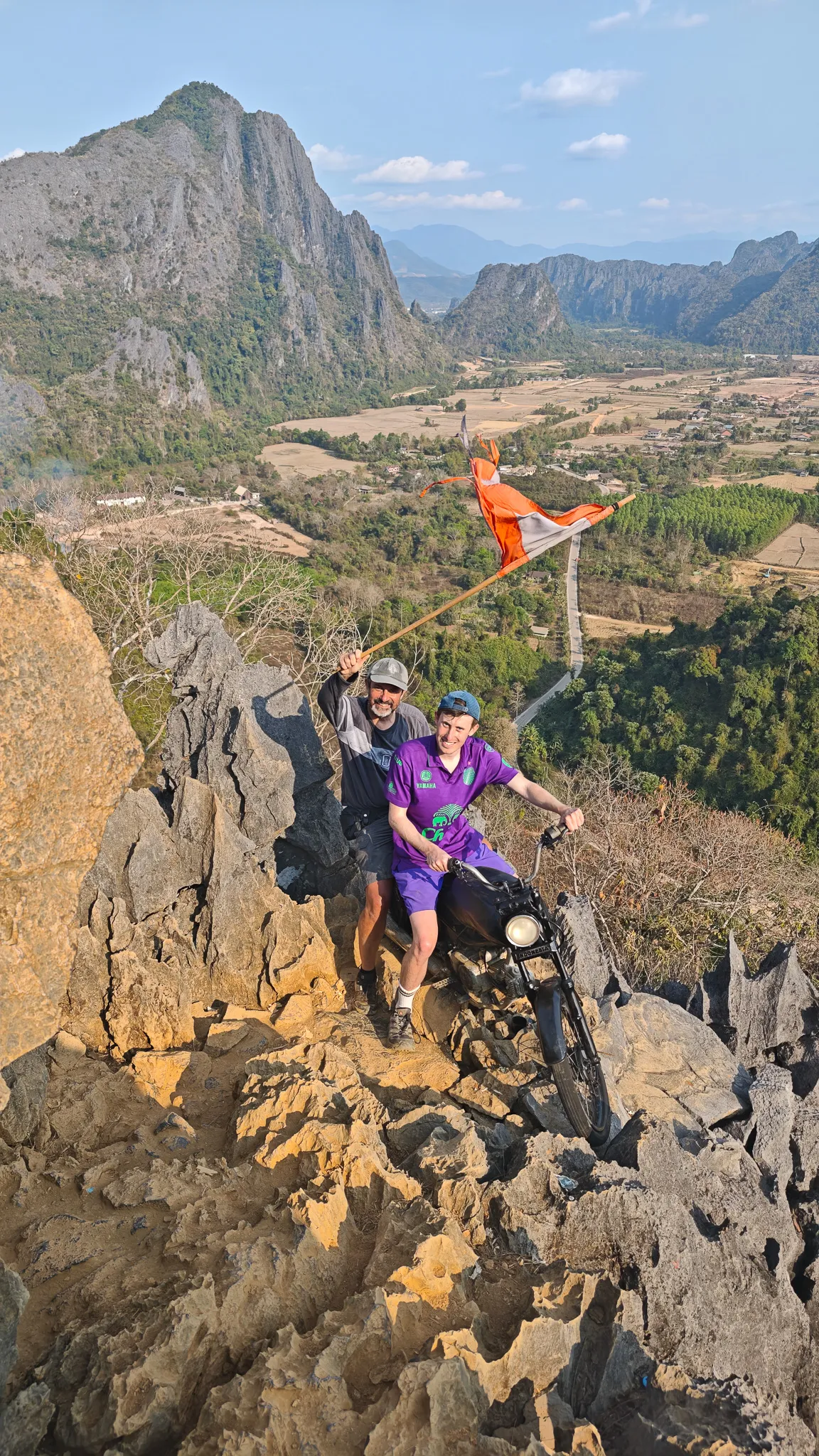 Two friends posing together at the Nam Xay summit next to the Laos flag and a motorcycle on the peak
