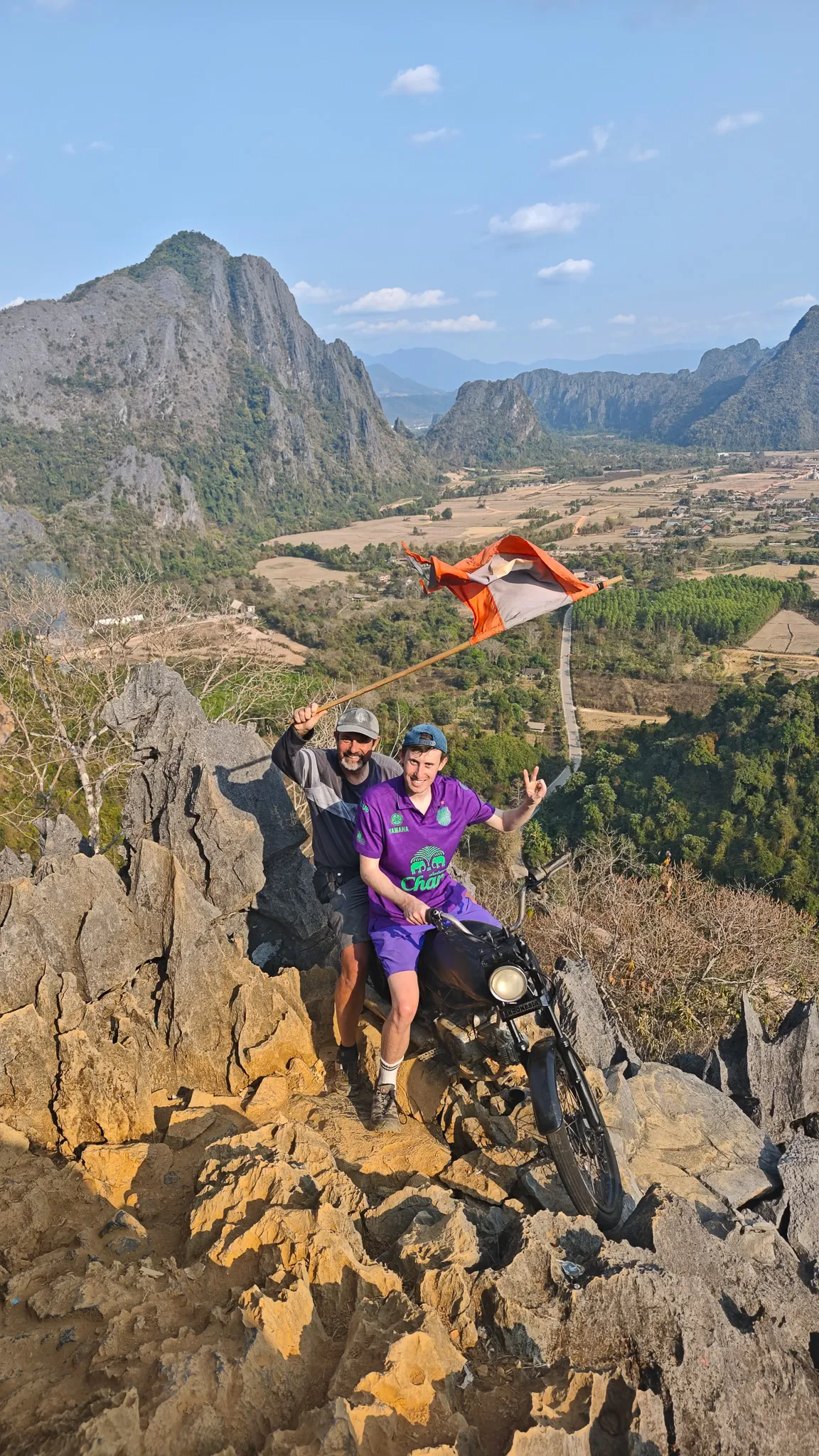 Selfie with a motorcycle and Laos flag at the Nam Xay viewpoint summit with mountains and valley views