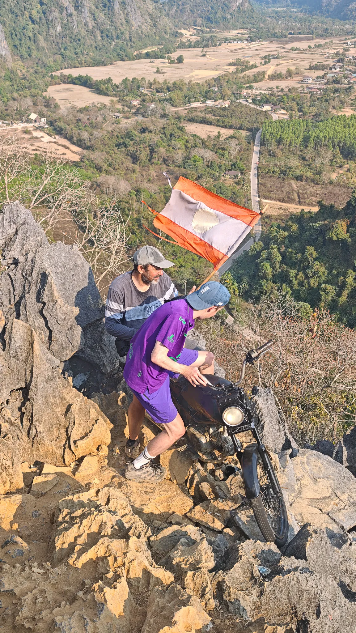 Two friends sitting at the summit of Nam Xay viewpoint with the Laos flag and karst mountains behind