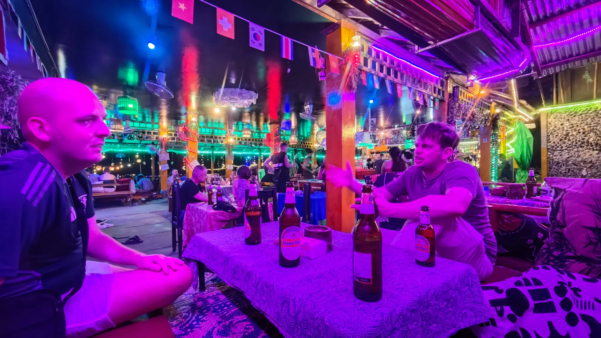 Two friends drinking Beer Lao at a neon purple-lit bar with international flags in Vang Vieng