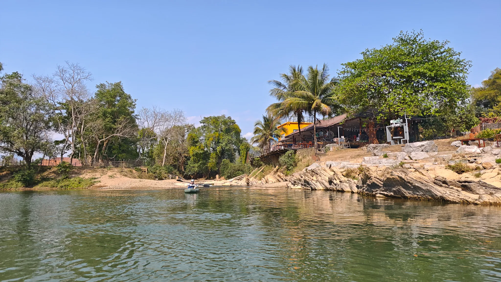 Packrafter paddling toward a riverside bar with palm trees along the Nam Song River in Vang Vieng