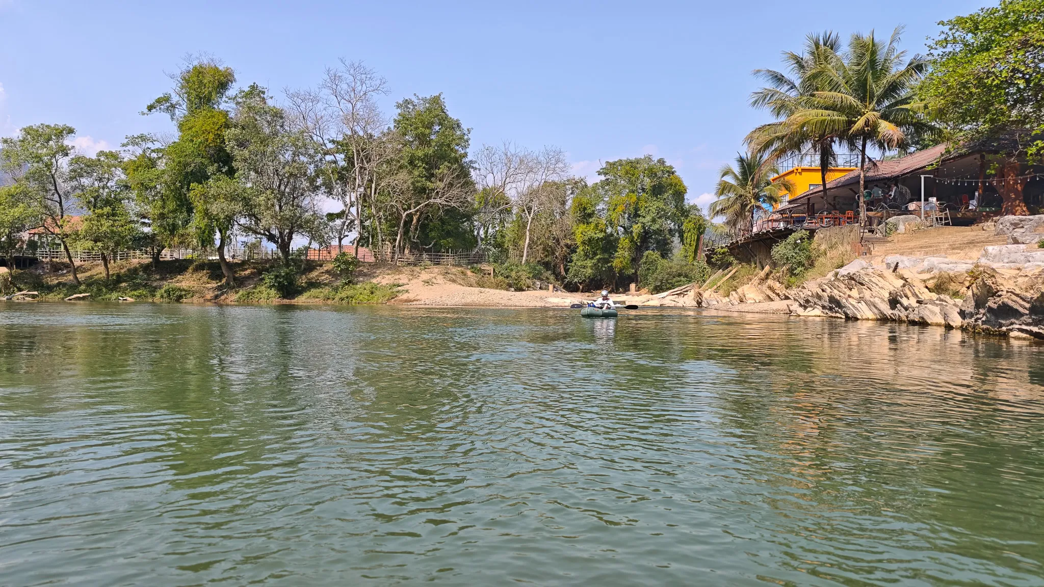 Distant packrafter on the Nam Song River near palm trees and a riverside bar in Vang Vieng