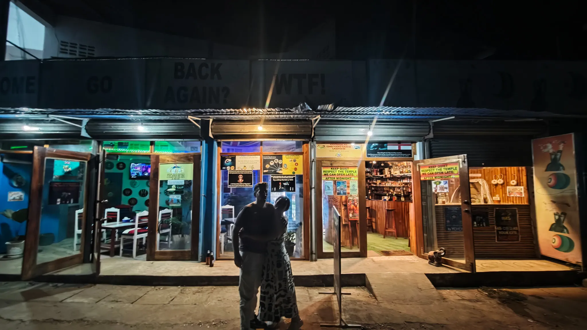 Exterior of a secret dive bar at night in Vang Vieng with neon signs reading Back Again and WTF