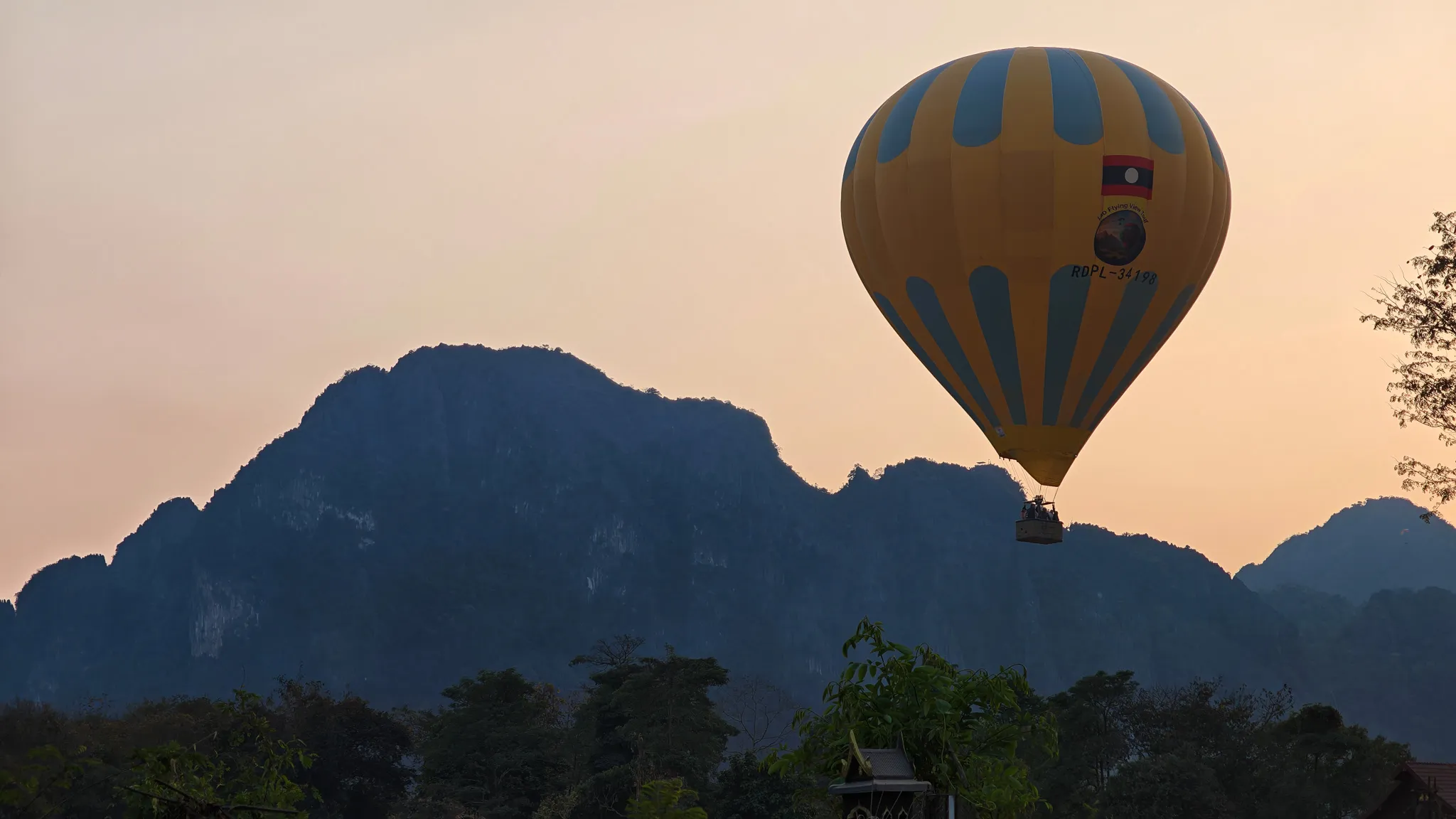 Yellow and blue hot air balloon with Laos flag floating above silhouetted karst mountains at sunset in Vang Vieng